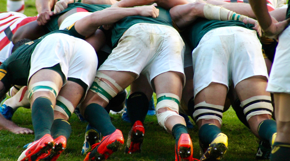 Rugby players contesting a scrum. Players in green socks and white shorts with their backs to camera, opposition players in red and white hooped shirts.
